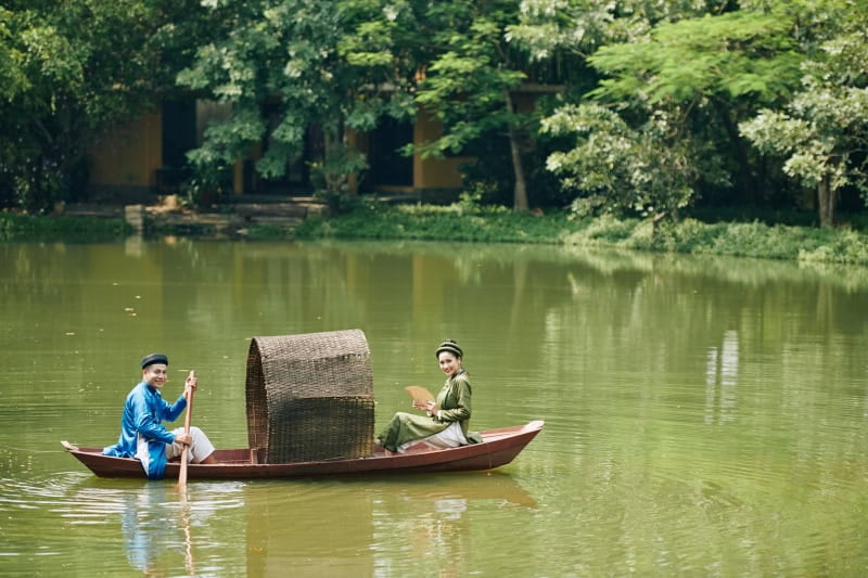 Smiling just married Vietnamese couple in traditional dresses and headwear rowing boat in small pond